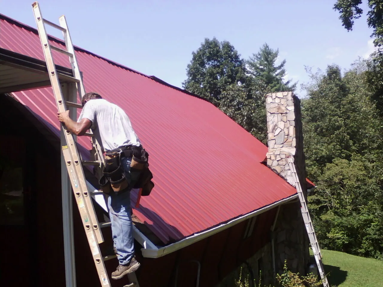 Roofer on a ladder beside a red metal roof and stone chimney — Soffit & Fascia Repair by Roofing Brothers in Pinecliffe
